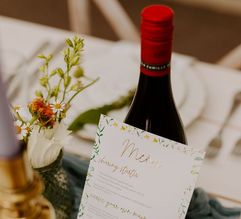 DIY wedding menu with gold foil leaning against red wine bottle 