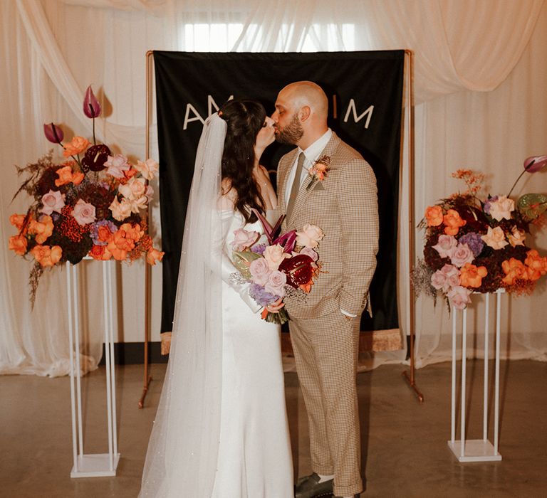 The bride and groom stand in front of the DIY wedding banner sign with drapery and colourful flowers 