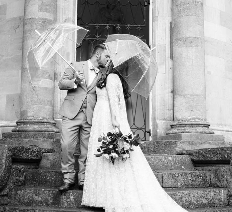 The bride and groom share a kiss as they hold umbrellas on the rainy wedding day 