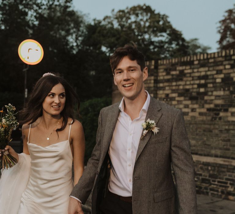 Groom in checked blazer and bride in slip wedding dress crossing the road at dusk 