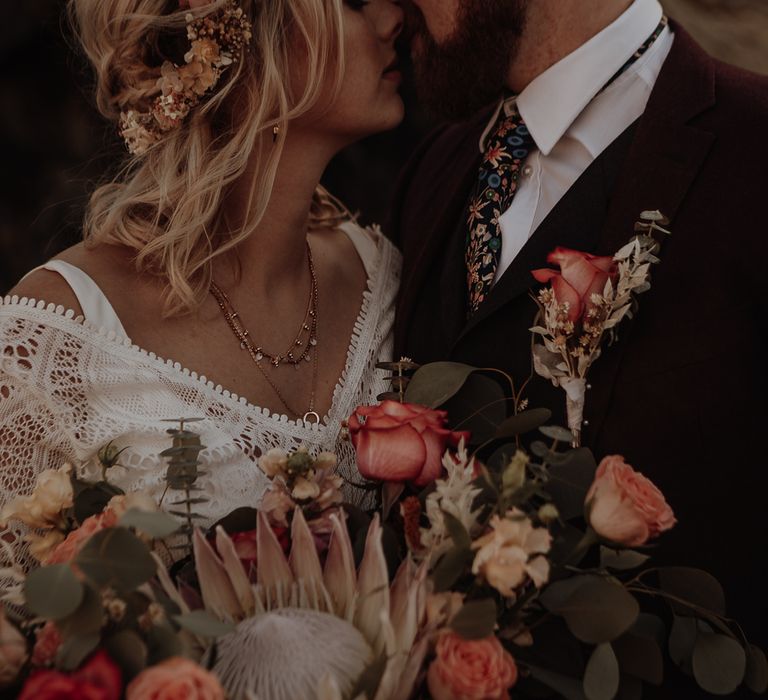 Groom with floral patterned tie and rose boutonnière next to bride with flower crown and gold necklaces holding a large bouquet of assorted flowers 