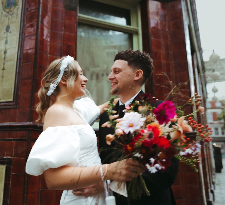 Bride and groom celebrate in the streets of London with a ruched off the shoulder wedding dress and colourful bouquet