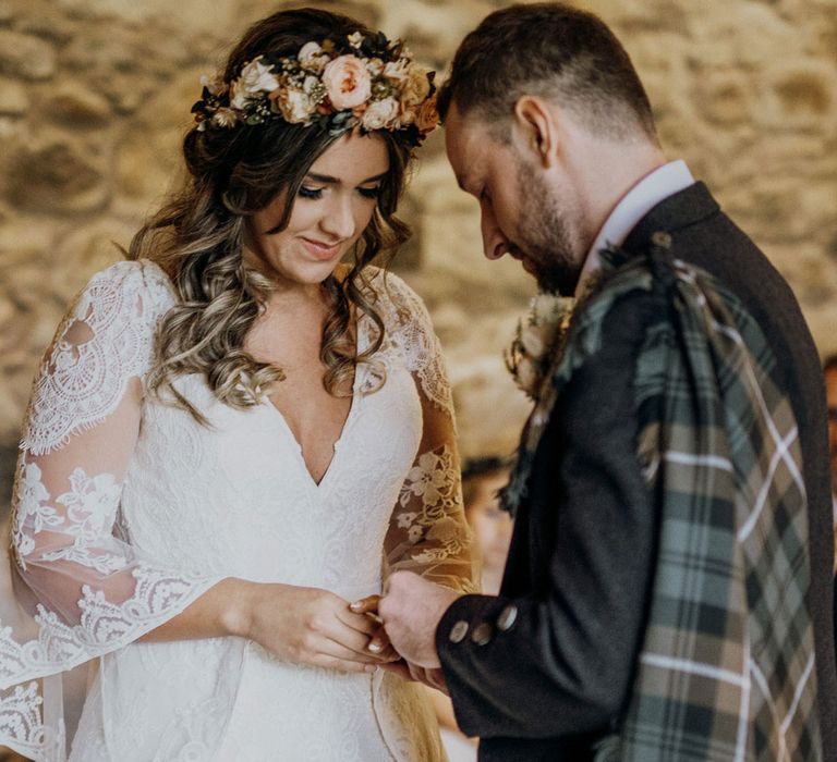 Groom in tartan kilt puts ring on the finger of boho bride wearing pink rose flower crown 