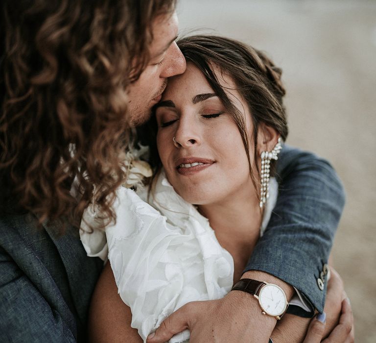 Bride with a nose ring and chandelier earrings in a ruffle sleeve wedding dress being kissed on the head by her curly hair groom