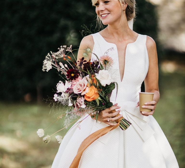 Beautiful blonde bride in a Jesus Peiro wedding dress with waffle fabric, plunging neckline and bow waist holding a wildflower bouquet tied with ribbon