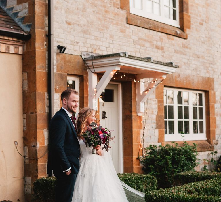 Bride & groom laugh and smile outside house whilst bride holds pink floral bouquet