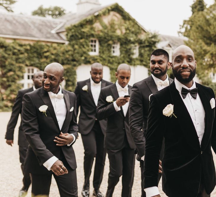 A groom walks with his groomsmen to his wedding ceremony. They wear black tuxedos.