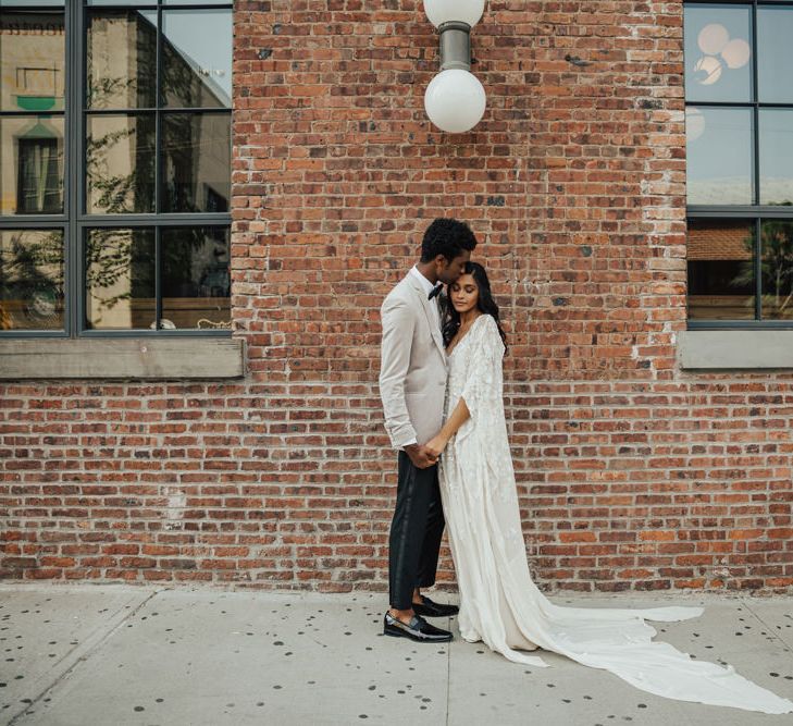 Groom in black tuxedo trousers with a grey blazer and bow tie standing with his bride in an embellished wedding dress with bat wing sleeves 