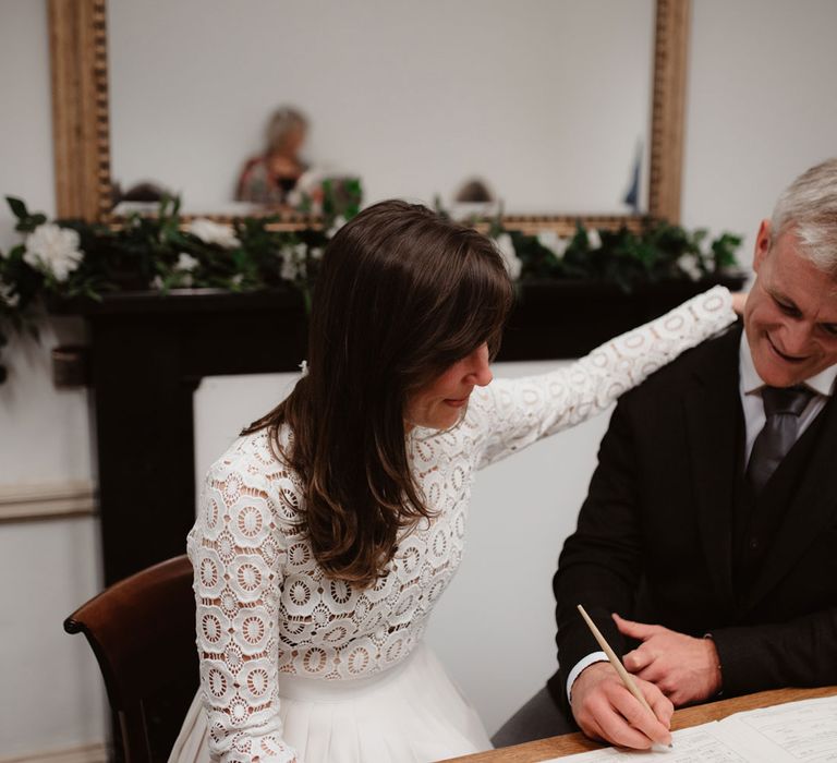 Bride in long sleeved lace top Self Portrait wedding dress holds shoulder of groom in brown woollen jacket and waistcoat as they sign marriage register