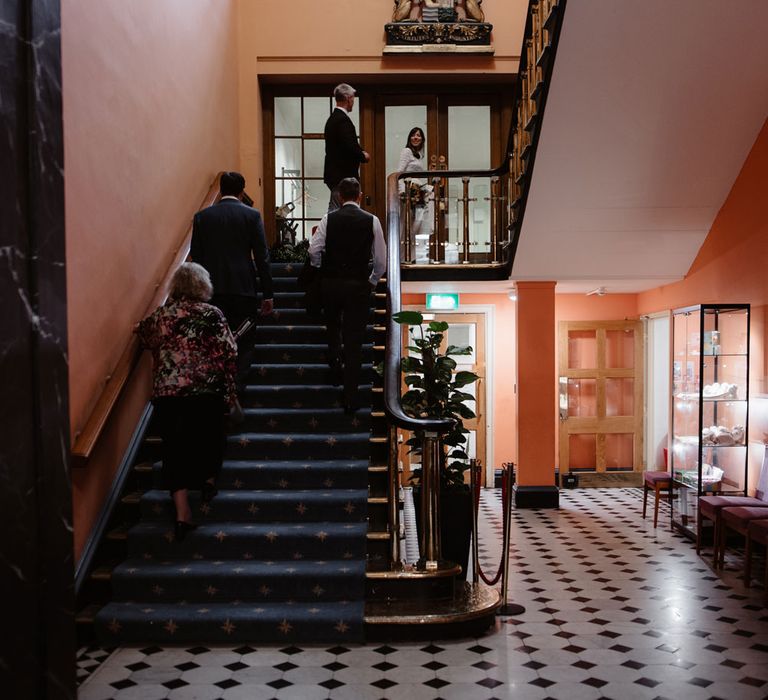 Bride in Self Portrait wedding dress, groom and witnesses walk up the stairs inside Bristol Registry Office