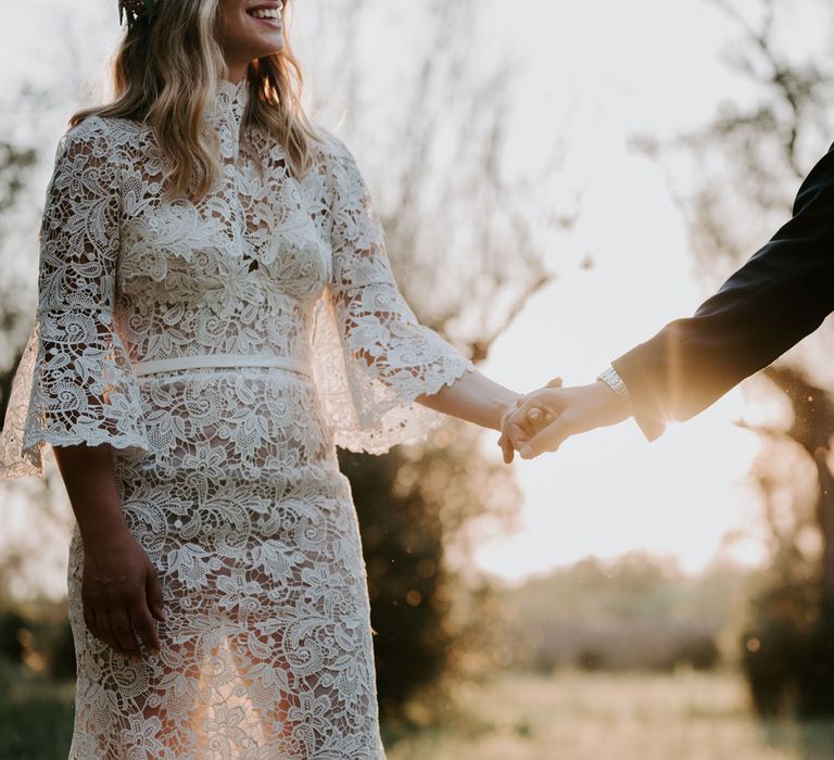 Bride in long sleeved lace boho wedding dress holds hands with groom in navy suit at enchanted forest wedding