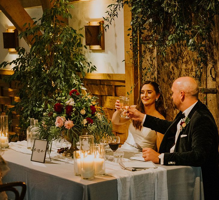 Bride and groom clinking glasses at their sweetheart table decorated with candles and foliage decor 