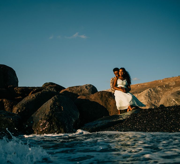 Groom sits behind bride on the rock as they watch the ocean 