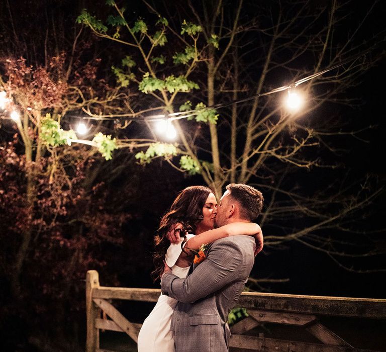 Bride in white strapless Vagabond wedding dress with train kisses groom in grey Hugo Boss suit and brown shoes outside at lakeside wedding