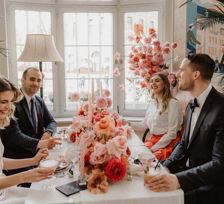 Bride, groom and two guests seated at the wedding table with cocktails and peach wedding flower centrepieces including pink, peach and burgundy ranunculi, garden roses and orchids