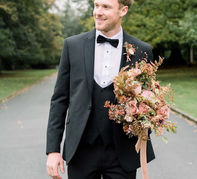 Groom in a black tuxedo and horseshoe waistcoat holding an autumn wedding bouquet tied with ribbon