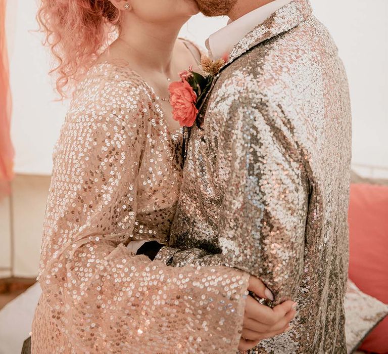 Bride with pink curled hair and beaded crown headband wearing gold sequin wedding dress kisses groom in silver sequin suit jacket in teepee at festival themed wedding 