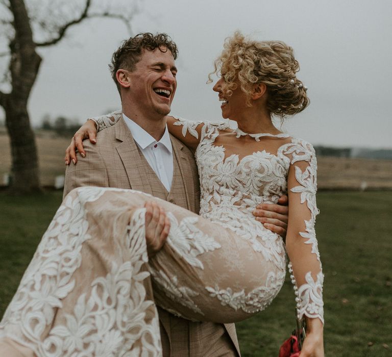 Groom carrying the bride and laughing as they walk through the fields of Hornington Manor