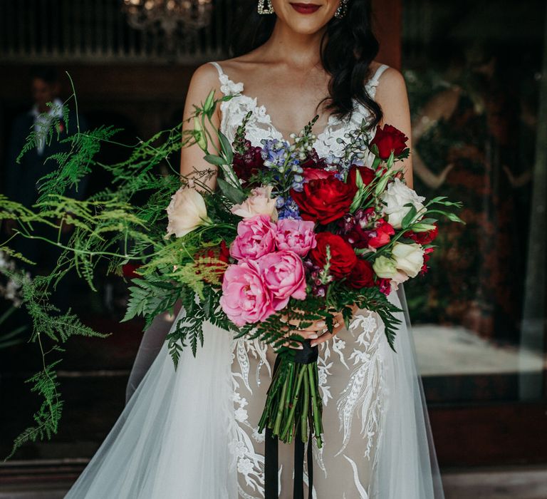 Bride carrying a large bouquet of red and pink peonies and roses with fern foliage