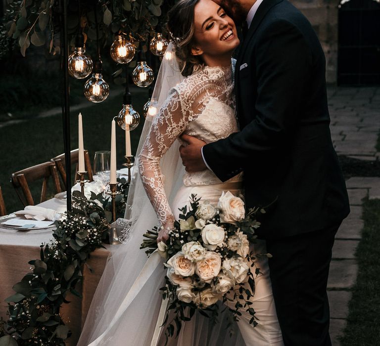 Groom in a black suit laughing with his bride in a long sleeve lace wedding dress as they sit on the edge of their elegant tablescape with festoon lights, greenery table runner and candles 