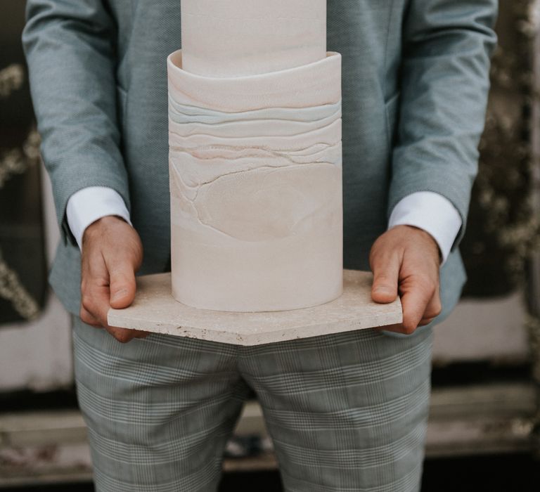 Groom holds wedding cake whilst wearing pastel blue suit 