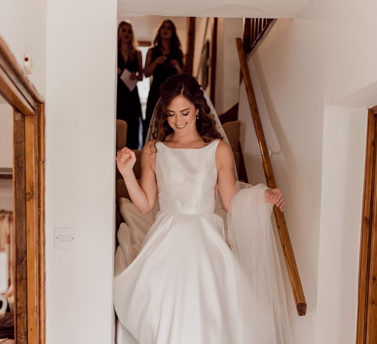 Bride in white satin Elbeth Gillis wedding gown walks down stairs inside house holding veil on one arm