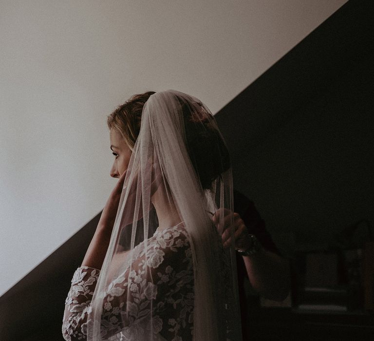 Bride in mesh floral embroidered top with three-quarter length sleeves looks out window whilst adjusting wedding veil before garden ceremony with burnt orange wedding theme