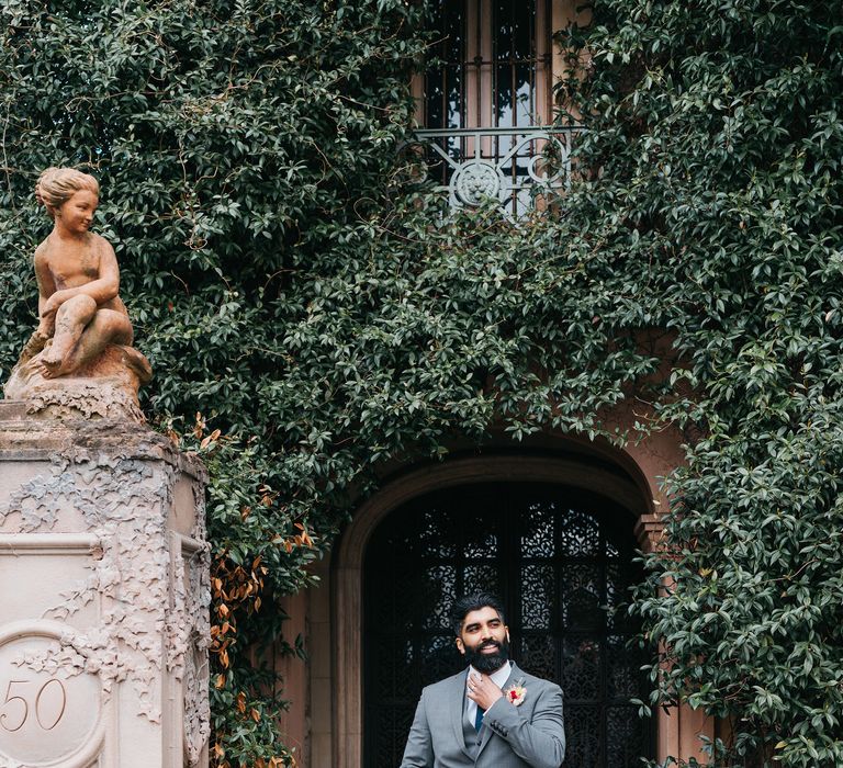 Groom stands within door way which is surrounded by green foliage as he wears Ted Baker suit and adjusts his tie