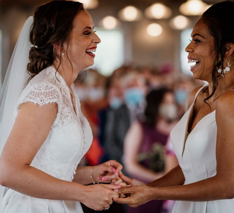 Laughing bride in white cami wedding dress with shoulder veils holds hands with smiling bride in lace capped sleeve wedding dress and veil in front of applauding guests during wedding ceremony at The West Mill Derby