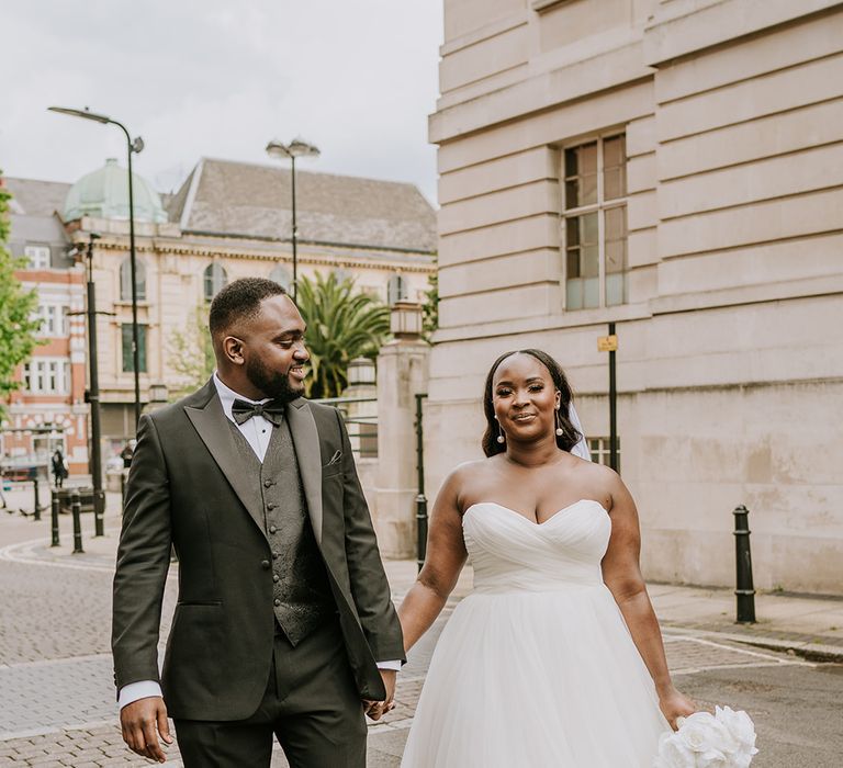 Bride & groom walk hand in hand on their wedding day outdoors as bride holds white and elegant bridal bouquet