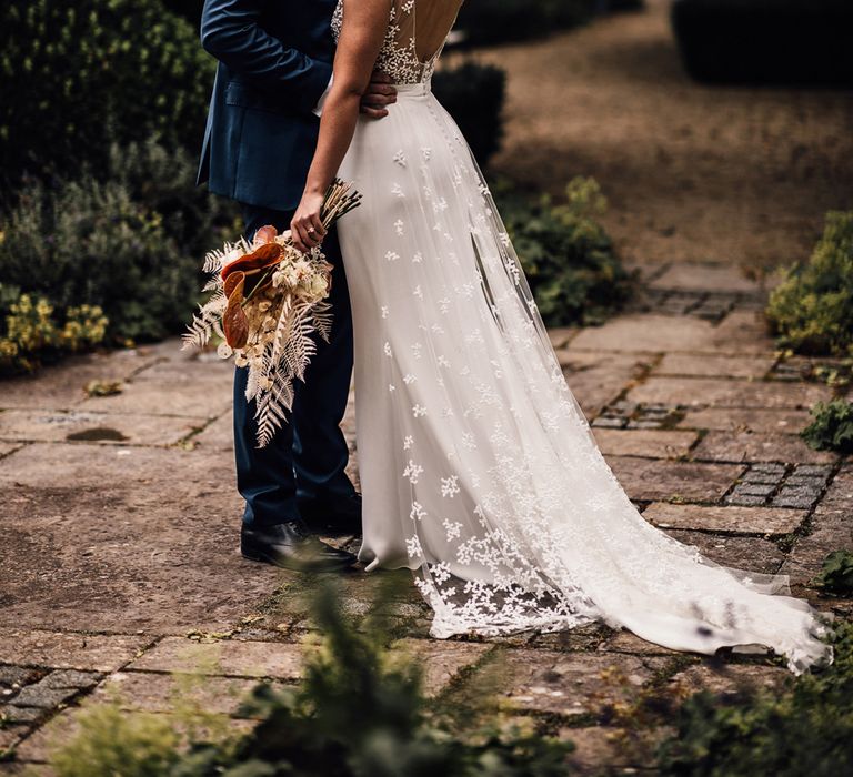 Bride in open back Rime Arodaky wedding dress with lace train holding dried bridal bouquet kisses groom in blue three piece suit outside after summer wedding ceremony in Dorset