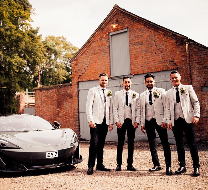 Stylish groomsmen in black trousers and pale grey jackets and horseshoe waistcoats standing next to a sports car 