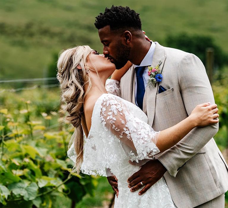 Groom in a beige suit kissing his bride in a lace wedding dress at Little Wold Vineyard