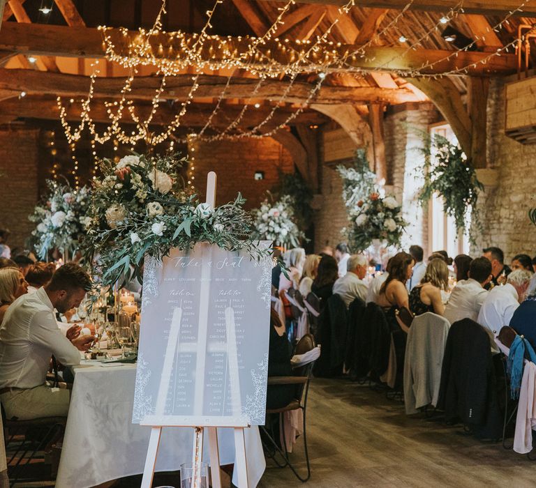 Guests sit at long white wedding tables in stone room with wooden beams lit by string fairy lights with frosted acrylic table seating plan wedding sign at Tythe Barn wedding with barn wedding flowers