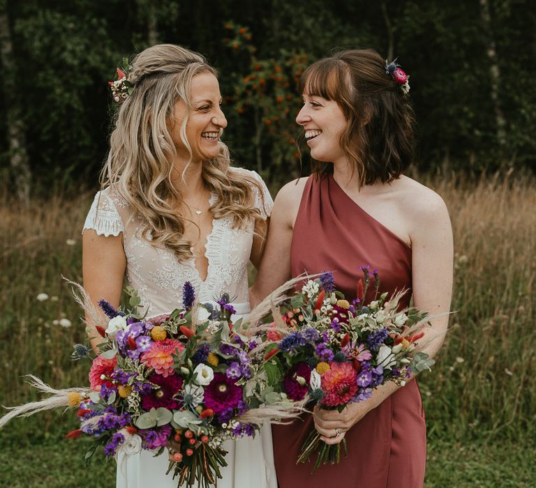 Bride in white lace top cap sleeved wedding dress and flowers in her hair smiles at bridesmaid in one shoulder burgundy bridesmaid dress as they both hold colourful mixed wedding bouquets during late summer wedding at Wellington Wood Norfolk