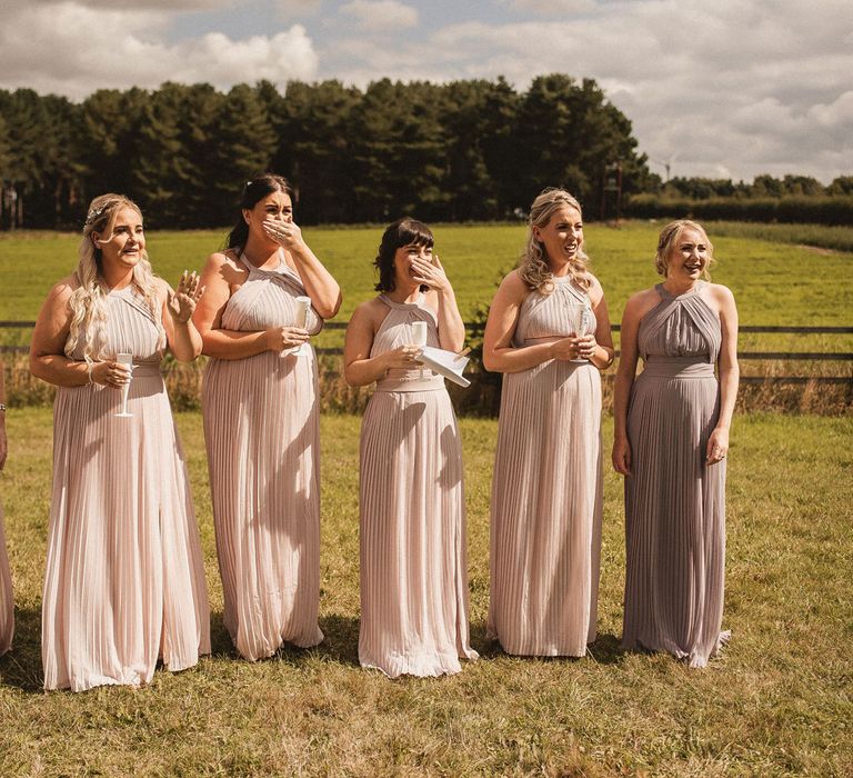 Bridesmaids in pink and purple bridesmaid dresses holding glasses of sparking wine have first look at the bride before tipi wedding at Inkersall Grange Farm