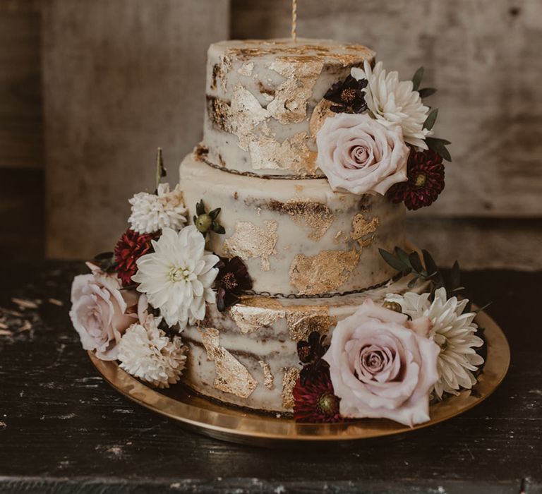 Semi naked wedding cake with gold leaf decor and flowers and a gold wire wedding cake topper of the couples names 