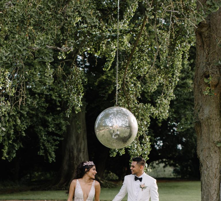 Bride and groom stand under large disco ball suspended from tree