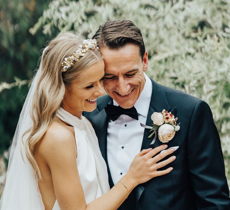 Bride in halterneck Halfpenny London wedding dress and veil wears bespoke Victoria Percival headband and hugs groom in black tie with rose floral buttonhole