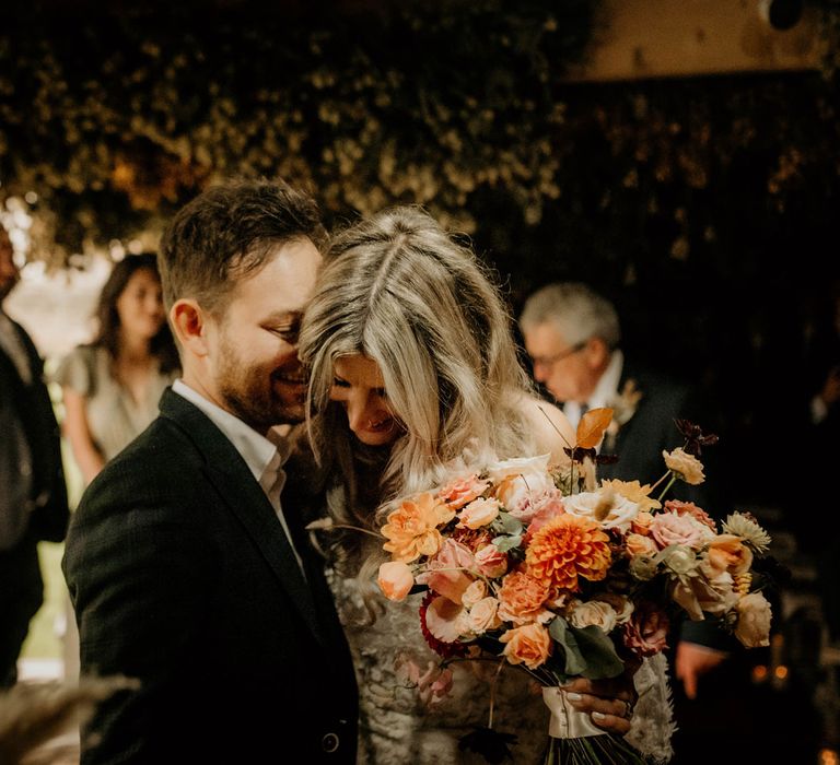 Smiling bride holding orange, pink and white autumnal bridal bouquet hugs groom in dark suit during rustic wedding ceremony