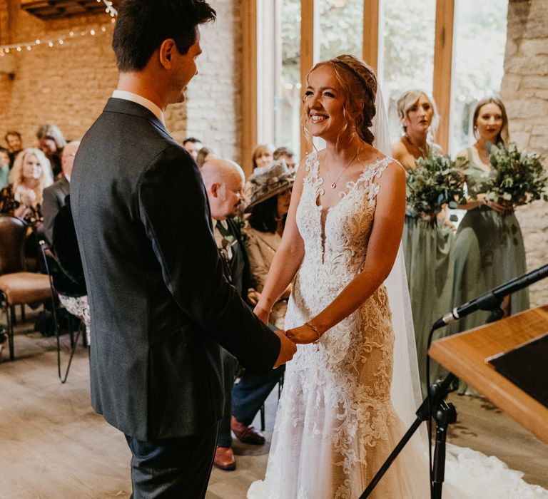 Bride in lace and tulle wedding dress stands holding hands with groom in dark suit during barn wedding ceremony