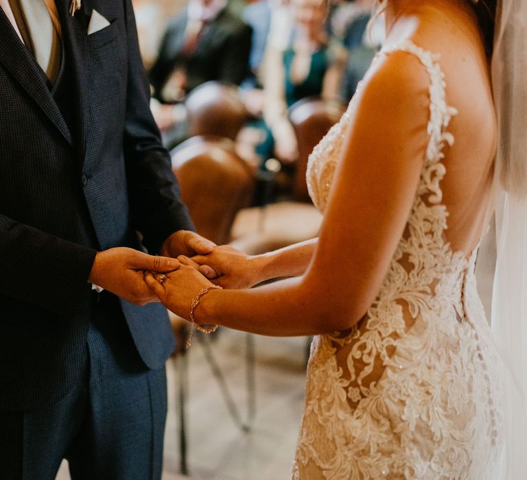 Bride in open back lace wedding dress and veil wearing bracelet holds hands with groom in dark three piece suit during wedding ceremony