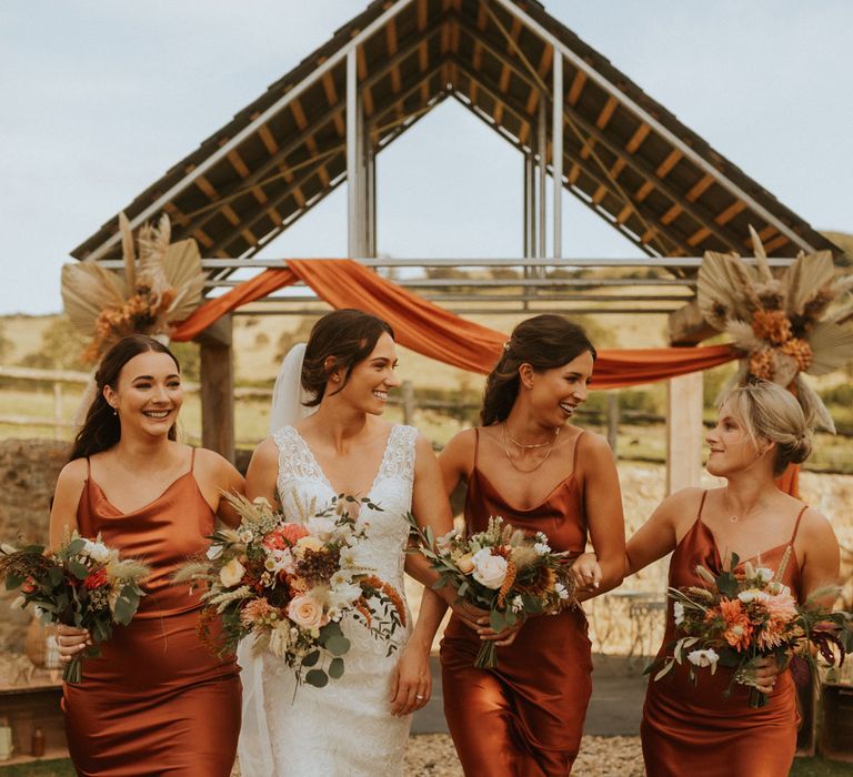 Bride in v neck lace wedding dress walks with bridesmaids in orange satin bridesmaid dresses all holding multicoloured bridal bouquets for rustic farm wedding