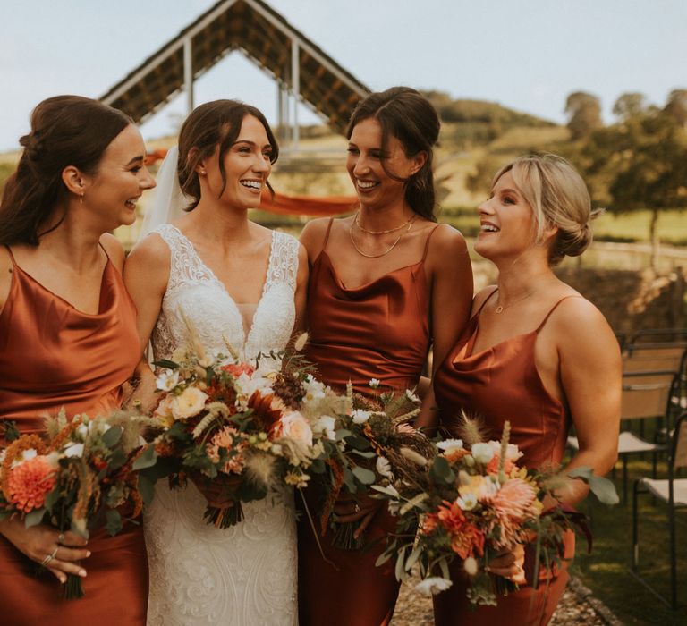 Bride in v neck lace wedding dress stands with bridesmaids in orange bridesmaid dresses holding mixed bridal bouquets at Hope Farm in Dorset