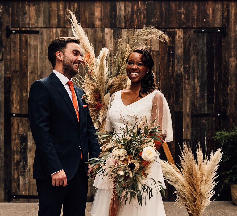 Black bride in bridal separates and groom in a navy suit standing at their wooden frame altar decorated with pampas grass and giant bottles 