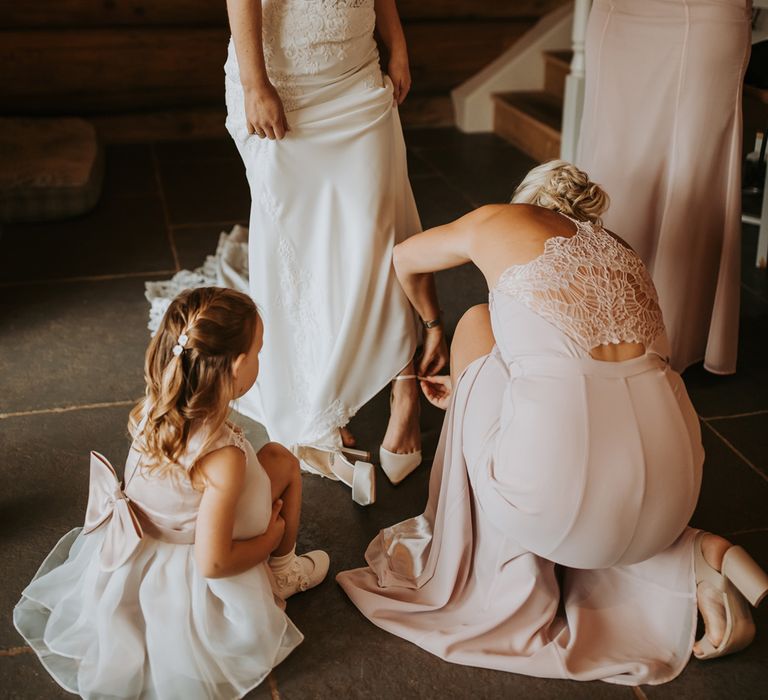 Bride with curled blonde hair and lace wedding dress stands whilst bridesmaids in pink bridesmaid dresses help her put on her shoes before wedding