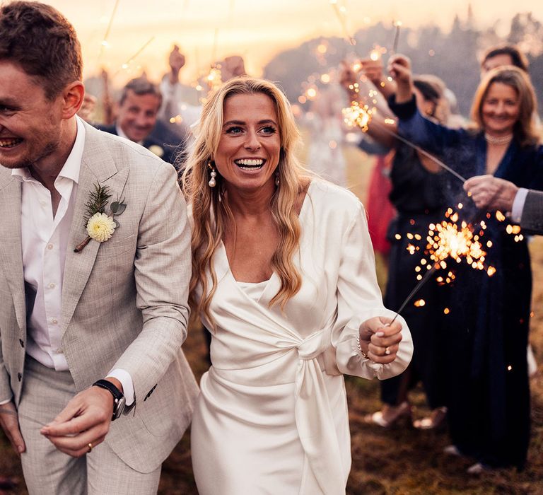 Smiling bride with drop pearl earrings and long sleeve satin wrap top walks with groom in grey suit with yellow floral buttonhole both holding sparklers