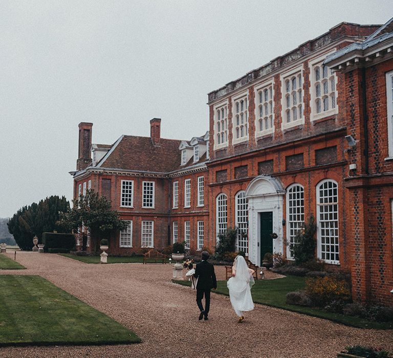 Bride & groom walk in front of Gosfield Hall on their wedding day 