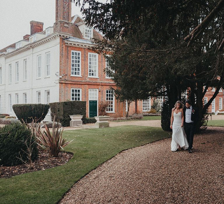 Bride & groom walk across gravelled pathway in front of Gosfield Hall 