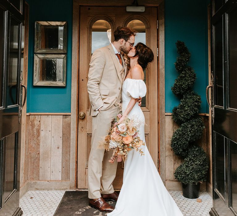 Groom in three piece beige suit kisses bride outside wedding venue with bride in puff sleeve sweetheart wedding dress 